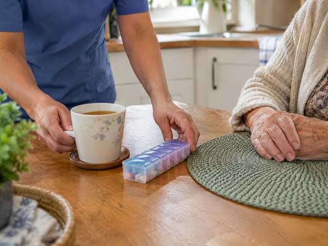 A home care worker's hands setting a cup of tea and a pill organizer in front of an older woman at a kitchen table
